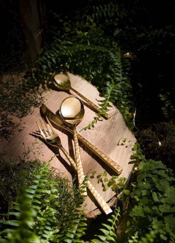 Gold Degrenne cutlery set displayed on a wooden surface surrounded by greenery